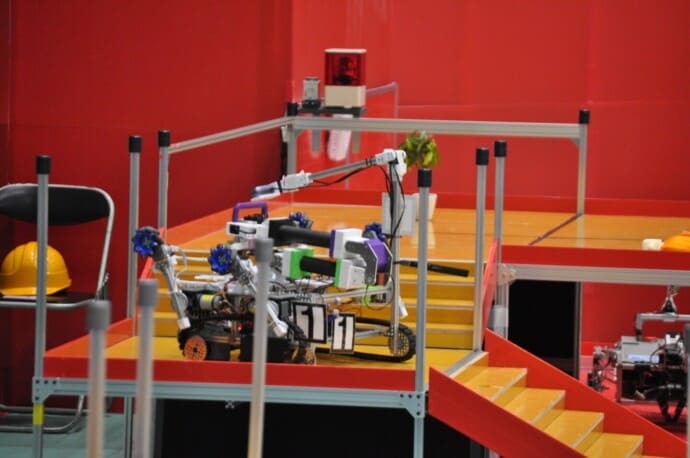 Robot with white and colored components on a yellow platform, numbered 11, inside a red-walled arena.
