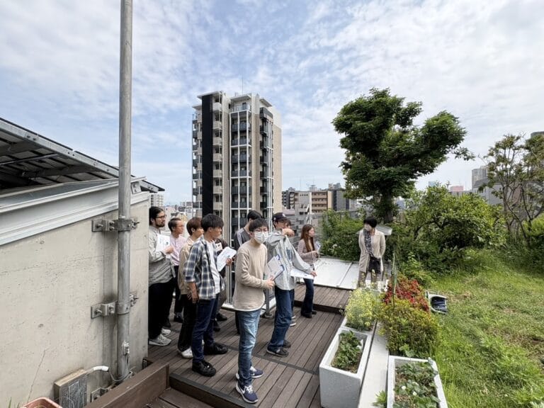 Group of people on a rooftop garden tour, holding papers and listening to a guide near a row of planters.