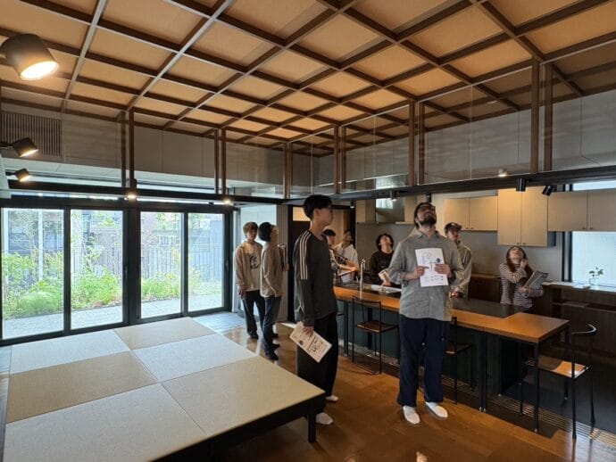Group of people indoors in a modern kitchen and lounge, some holding papers and looking up at the ceiling lights.