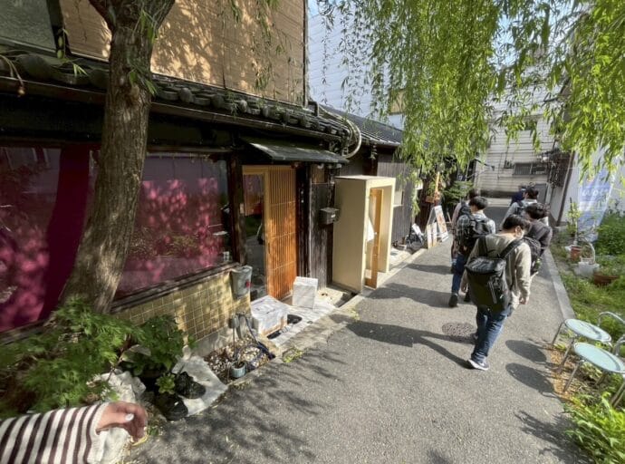 Narrow alley with a row of people wearing backpacks walking past a wooden storefront and greenery overhead.