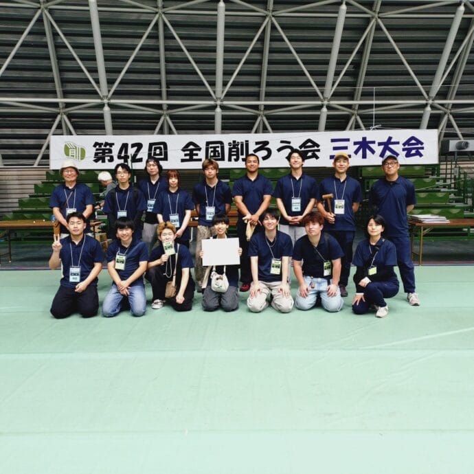 Group of people in blue shirts posing for a photo in an indoor stadium with a large white banner containing Japanese text behind them.