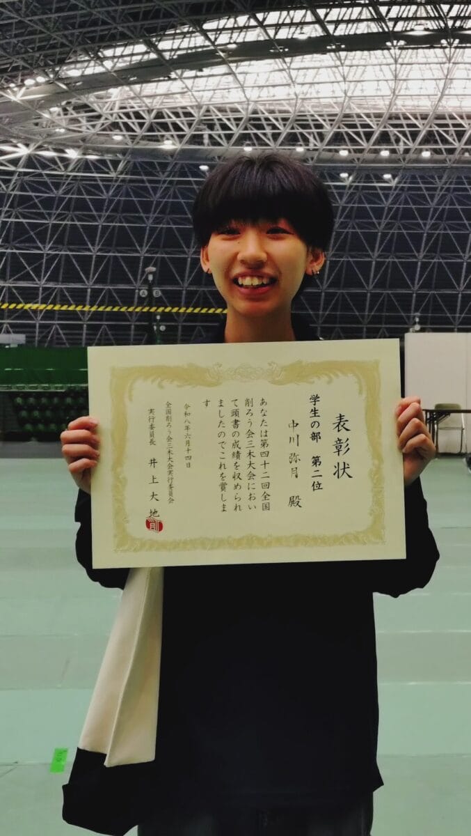 Young person smiling and holding a large Japanese certificate indoors, with a steel lattice roof in the background.