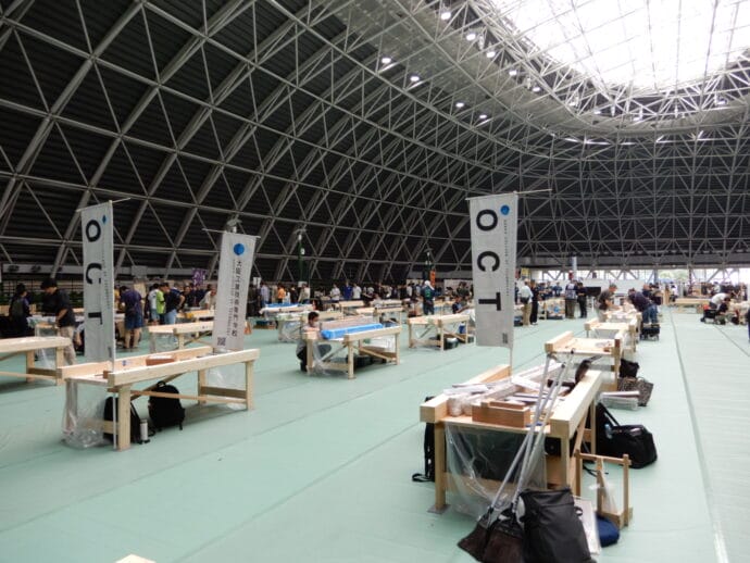 Wide view of a large indoor workshop hall with rows of workbenches and people at tasks under a lattice roof.