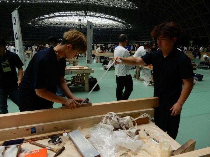 Two young men in dark shirts plane a wooden beam at a crowded woodworking event indoors.