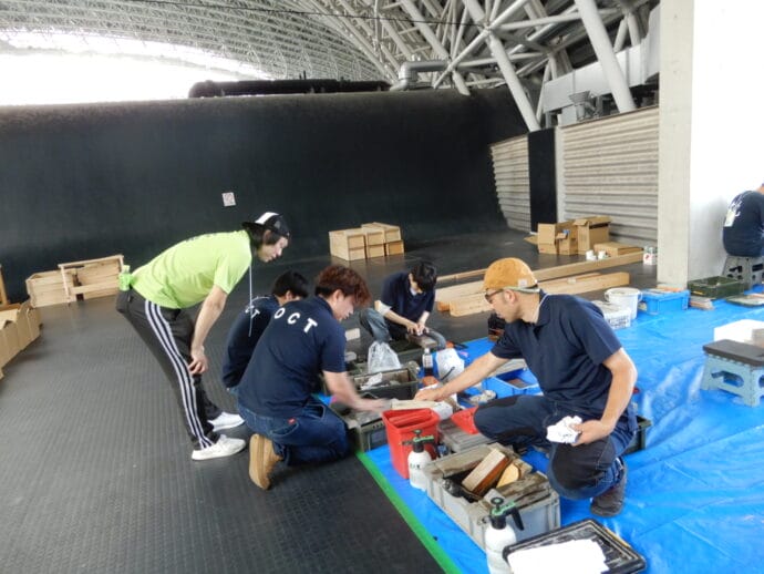 Group of people kneeling and working at a low workbench with wood pieces and tools in a large indoor workshop with a curved black wall behind them.