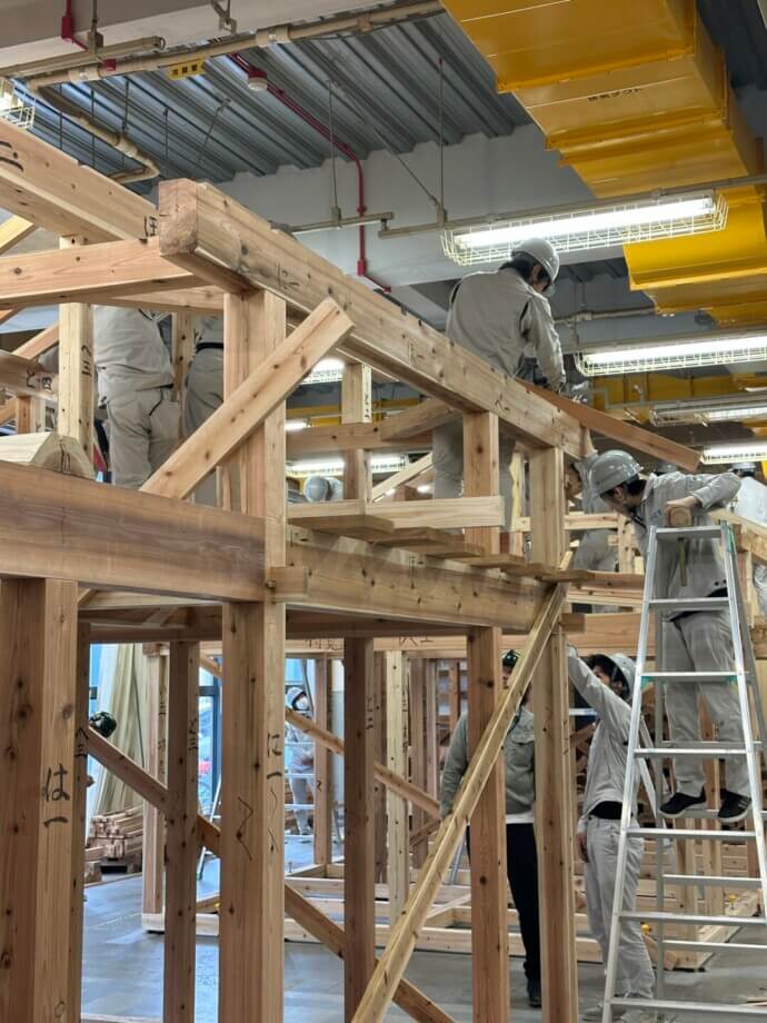Construction workers in hard hats assemble wooden framing on an indoor construction site with a ladder nearby.