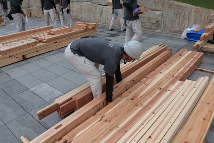 Worker wearing a hard hat and gray shirt bends over long wooden planks, organizing timber on a paved plaza area