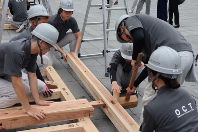 Group of workers in gray shirts and helmets assemble wooden beams on a paved plaza during a training exercise.
