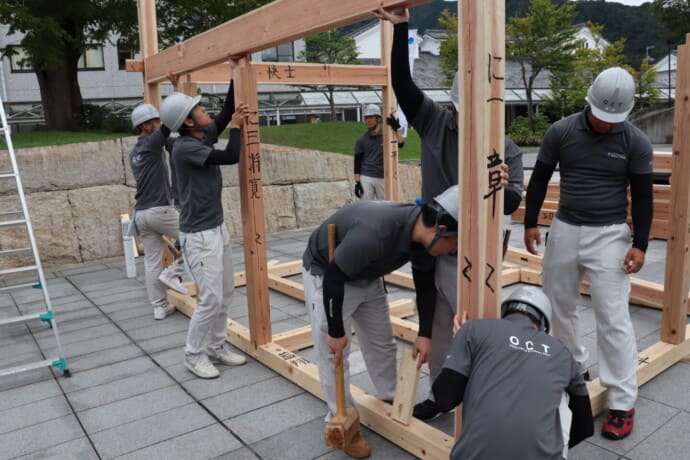 Construction workers in helmets assemble a wooden frame outdoors as part of a building project.