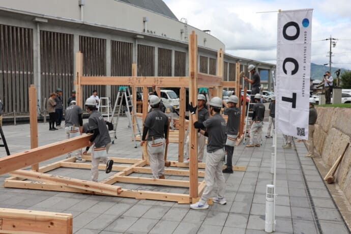Group of students in hard hats assembling a wooden frame outdoors with ladders and tools around them during a training event.
