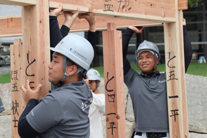 Three workers in helmets raise a wooden beam to form a frame, with handwritten Japanese characters on the posts in an outdoor construction site near a campus building.