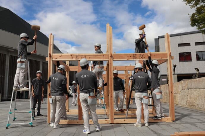 Construction crew assembling a wooden frame outdoors, workers in hard hats and tool belts coordinating on ladders and beams under a blue sky.