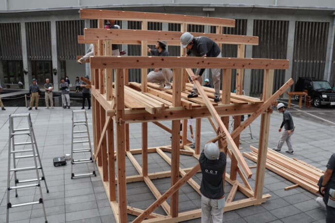 Team of construction workers in hard hats assembling a timber-framed structure outdoors in a paved courtyard, with ladders nearby