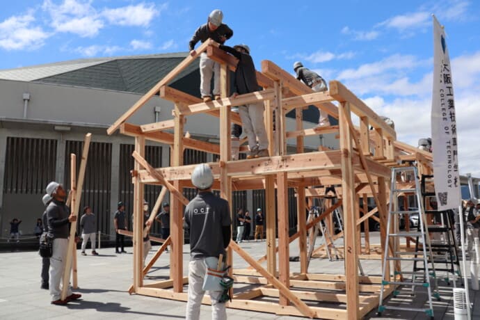 Outdoor construction site where workers in hard hats assemble a large wooden frame; ladders nearby and a blue sky above.