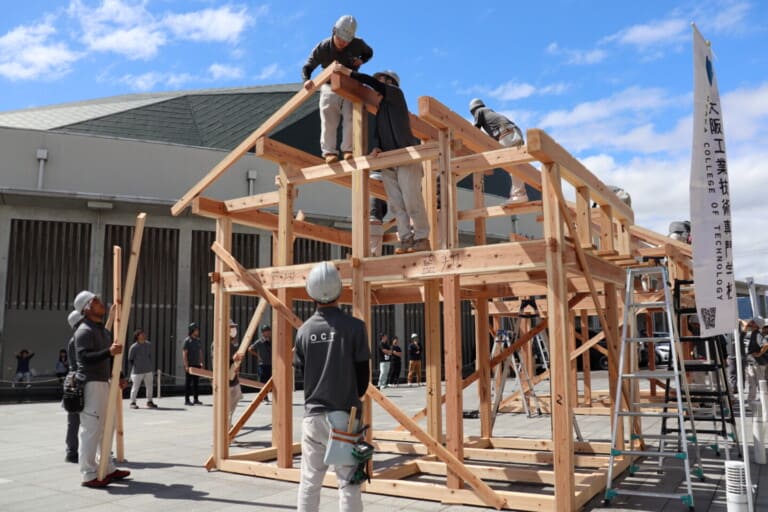 Outdoor construction site where workers in hard hats assemble a large wooden frame; ladders nearby and a blue sky above.