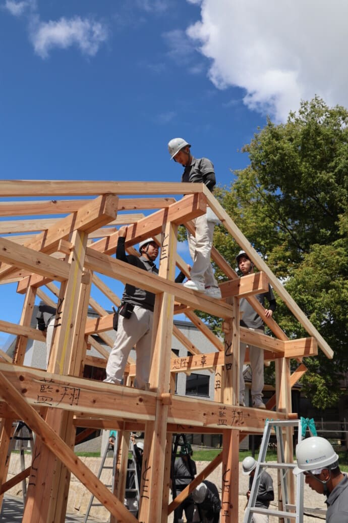 Construction workers wearing hard hats assemble a wooden frame against a clear blue sky.
