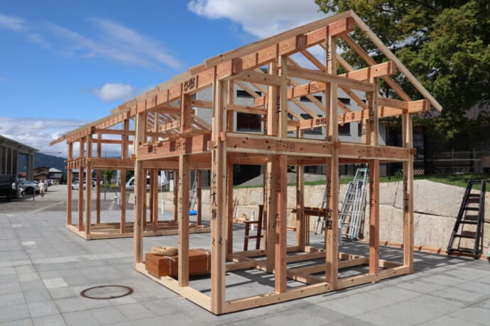 Open wooden house frame under construction in a paved public space, with ladders and a bright blue sky above.