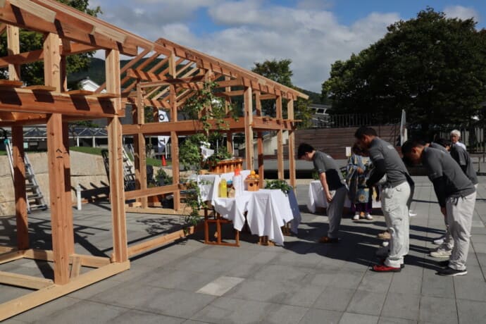 Group of people bowing before an offering table inside a wooden framing structure at an outdoor ceremony.