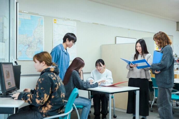 A diverse group of students works together around a table in a classroom, with maps on the wall and laptops open as they review papers.