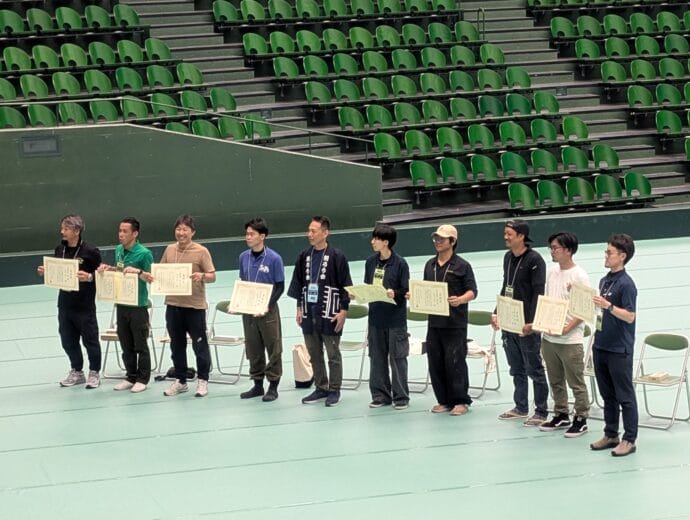Group of people standing in a gym, each holding a certificate, posing for a photo with green stadium seats in the background