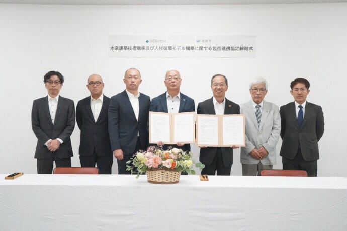 Seven men in business suits stand behind a long table as two of them hold open documents, during a formal signing ceremony with a flower centerpiece on the table and a banner overhead in Japanese text.
